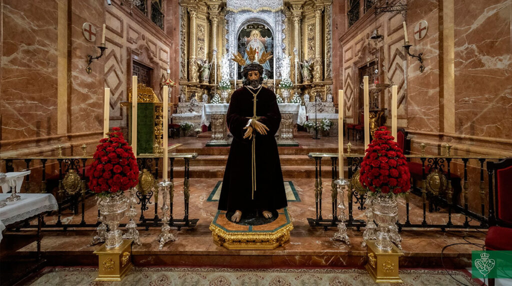 Nuestro Padre Jesús de la Sentencia presidiendo el Devoto Besamanos en la Basílica de la Esperanza Macarena, rodeado de centros de flores rojas y blandones.
